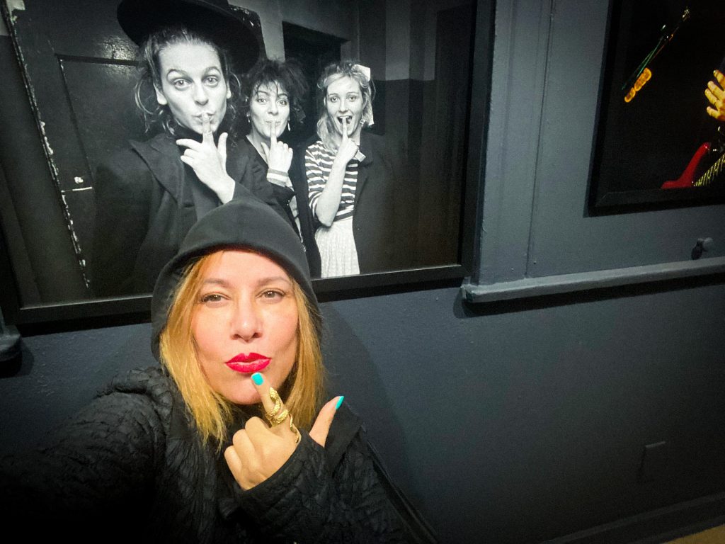 Jennifer Finch of L7 sits in front of a framed black and white photo of the punk band the Slits.