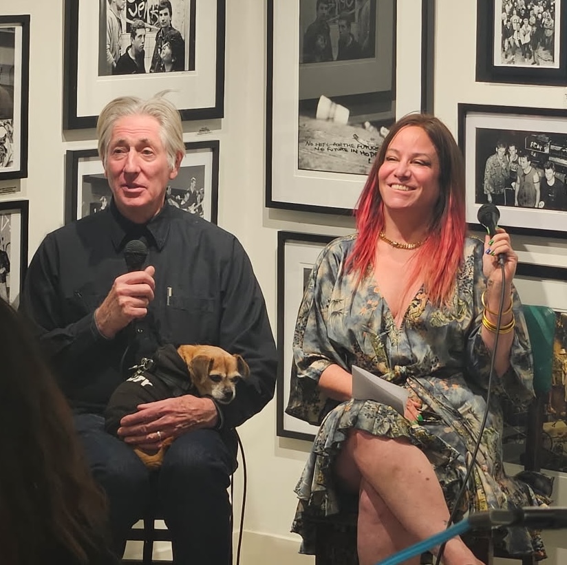 Jennifer Finch sitting with Edward Colver behind framed black and white photos of punk rock culture